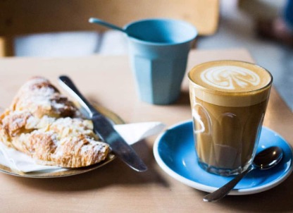 Photo of a coffee shop table. To the left of the image is an almond croissant. To the left is a flat white coffee in a clear glass with a blue saucer. A teaspoon lies on the saucer. 