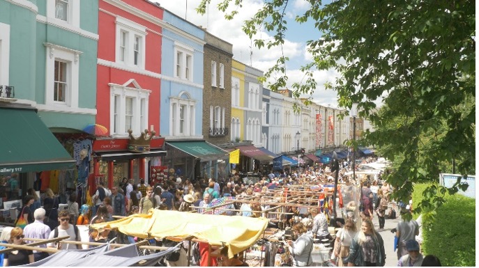 Photo of portobello road market. A line of colourful houses stand in front of stalls selling clothes. Crowds of people are standing around between the stalls. https://visitportobello.com/